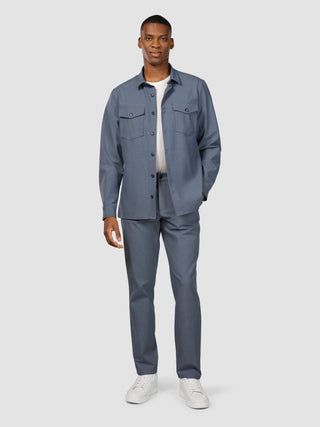 A man wearing a Storm Blue Melange Essential Overshirt over a white t-shirt, matching blue trousers, and white sneakers in a clean, Scandinavian design aesthetic.