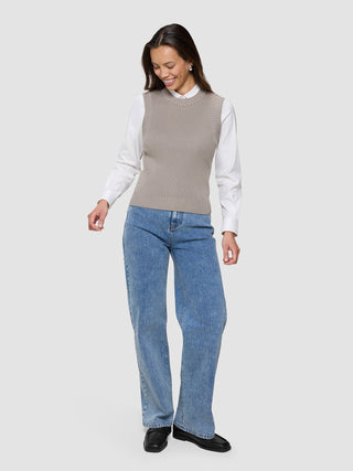 A smiling woman wearing a Women’s Fine Knit Rib Vest Dark Sand over a white button-down shirt, blue jeans, and black loafers.
