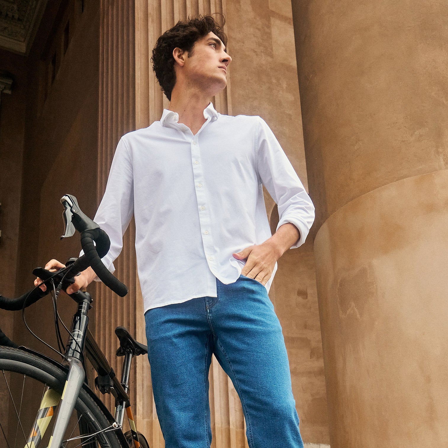 Man wearing white shirt and light blue jeans standing in front of an urban building with a bike