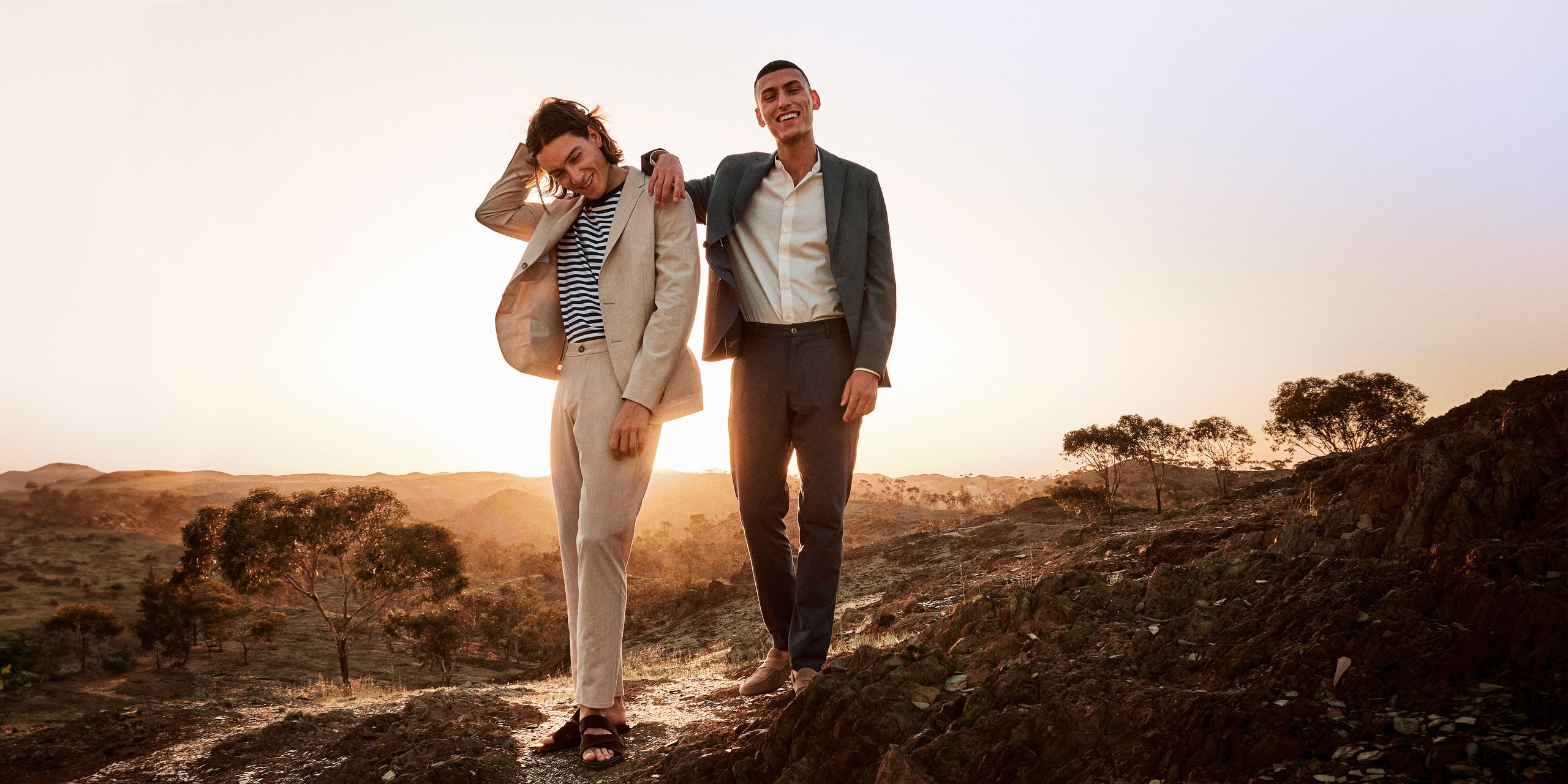 Two male models standing in a desert wearing a beige suit and grey suit looking happy