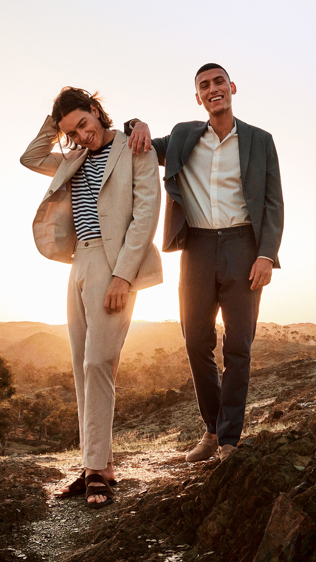 Two male models standing in a desert wearing a beige suit and grey suit looking happy