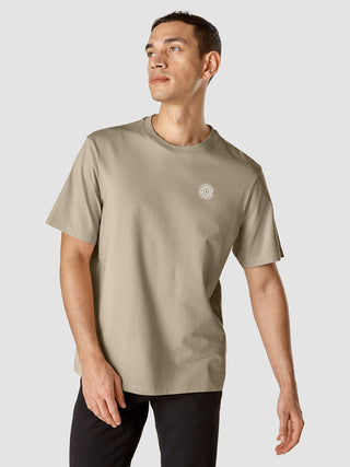 A man wearing a Sahara-colored Supima T-shirt Box Fit Globe Sahara with a small circular logo graphic on the chest.
