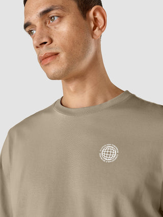 A man wearing a Supima T-shirt Box Fit Globe Sahara with a small circular logo on the chest featuring a globe and the text Shaping New Tomorrow Comfortable Anywhere.