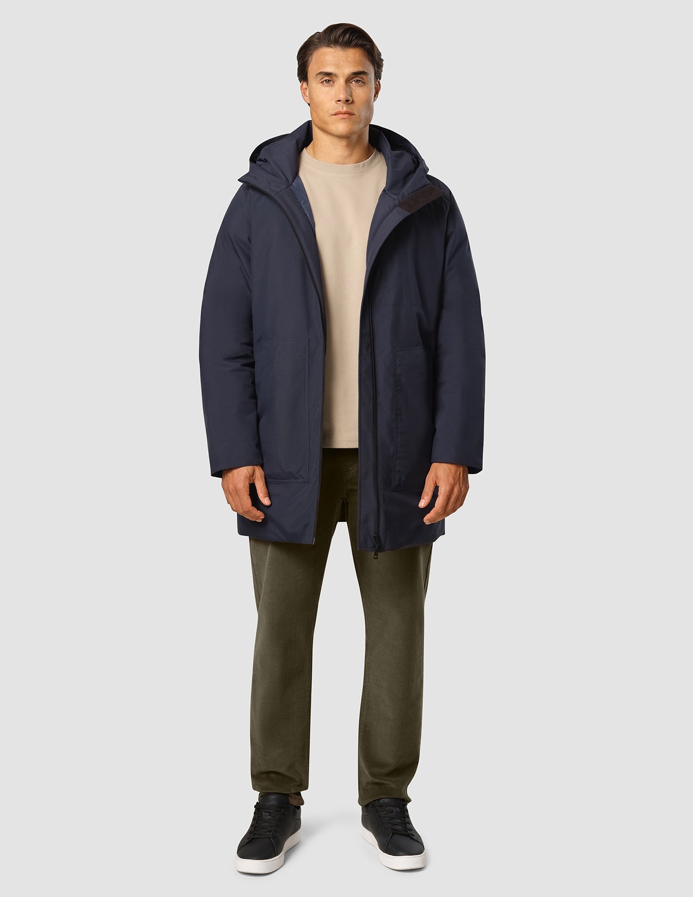 A man wears a Utility Jacket Dark Navy over a beige sweater, olive trousers, and black sneakers.