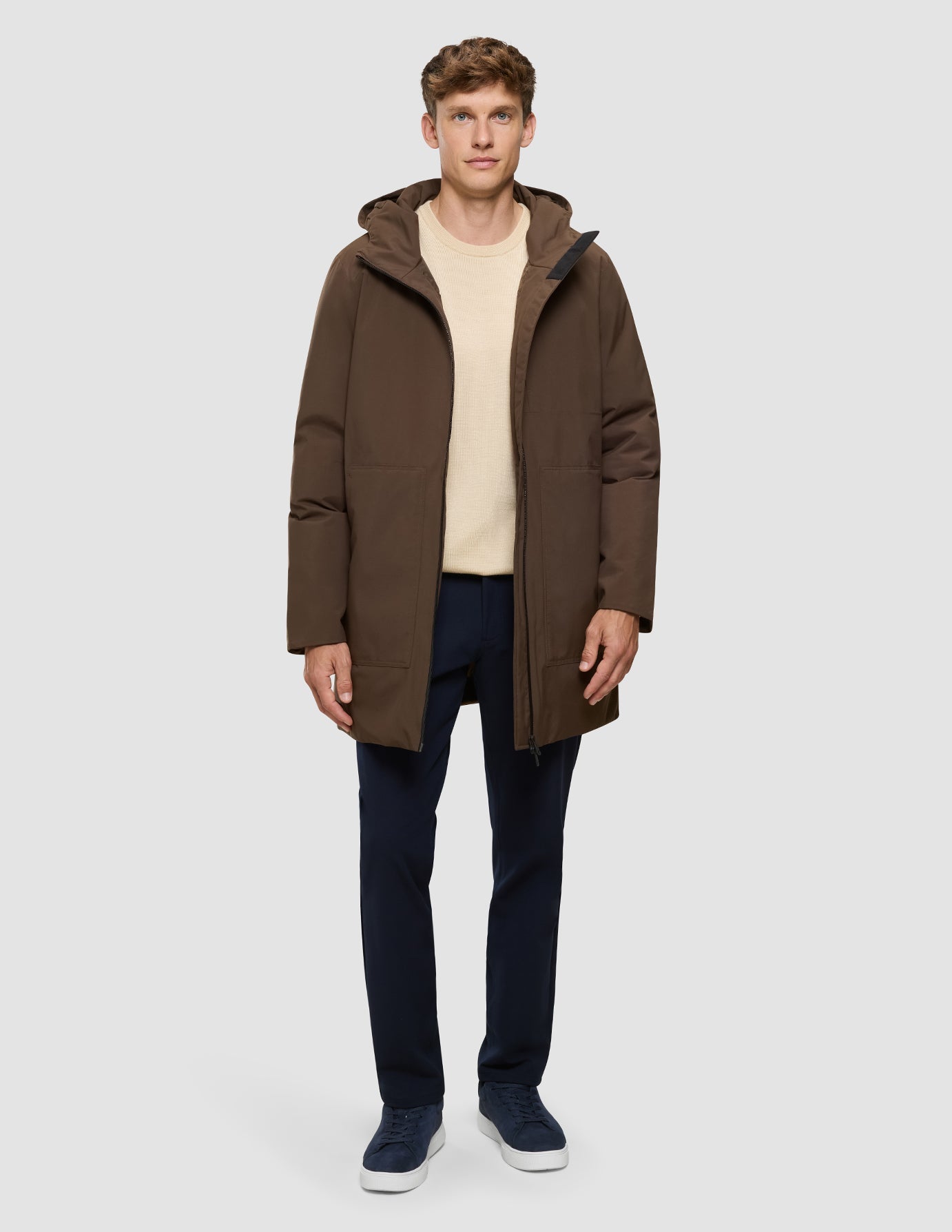 A man wearing a Utility Jacket Saddle Brown, a beige sweater, dark trousers, and dark sneakers.