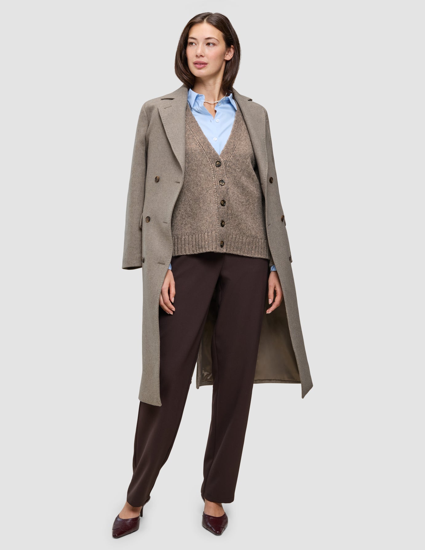 A woman wearing Cold-weather workwear consisting of a long beige wool coat over a brown cardigan, a light blue Oxford shirt, and dark trousers.