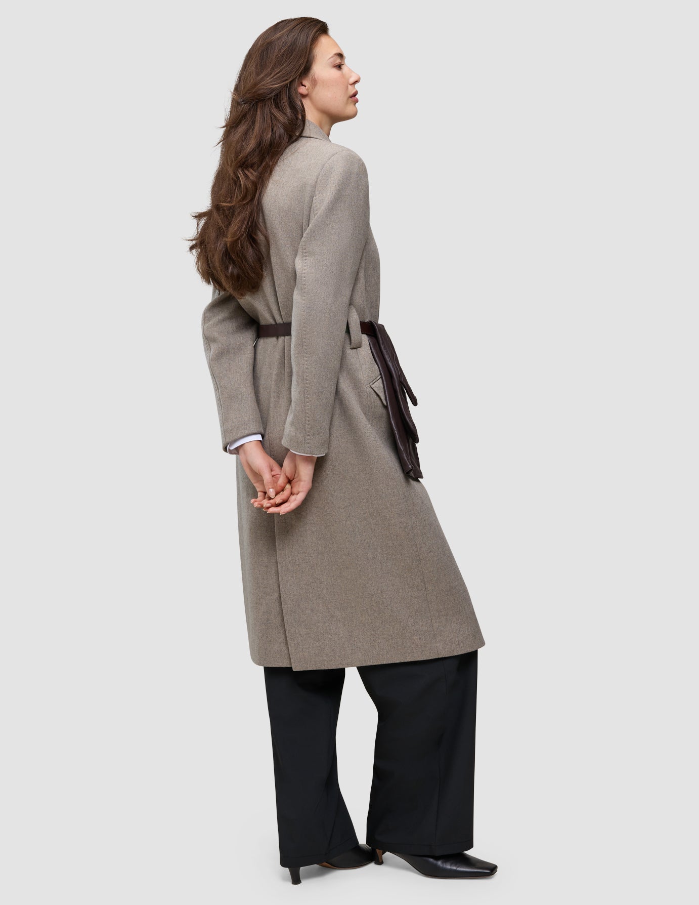 A woman seen from the side wears the Winter, dressed up look, featuring a long beige wool coat with a dark brown belt and black trousers.
