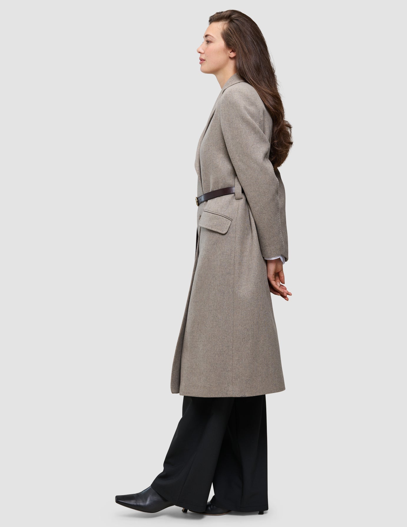 A person in profile wearing the Winter, dressed up beige wool coat cinched with a thin brown belt over an oxford shirt and black trousers.