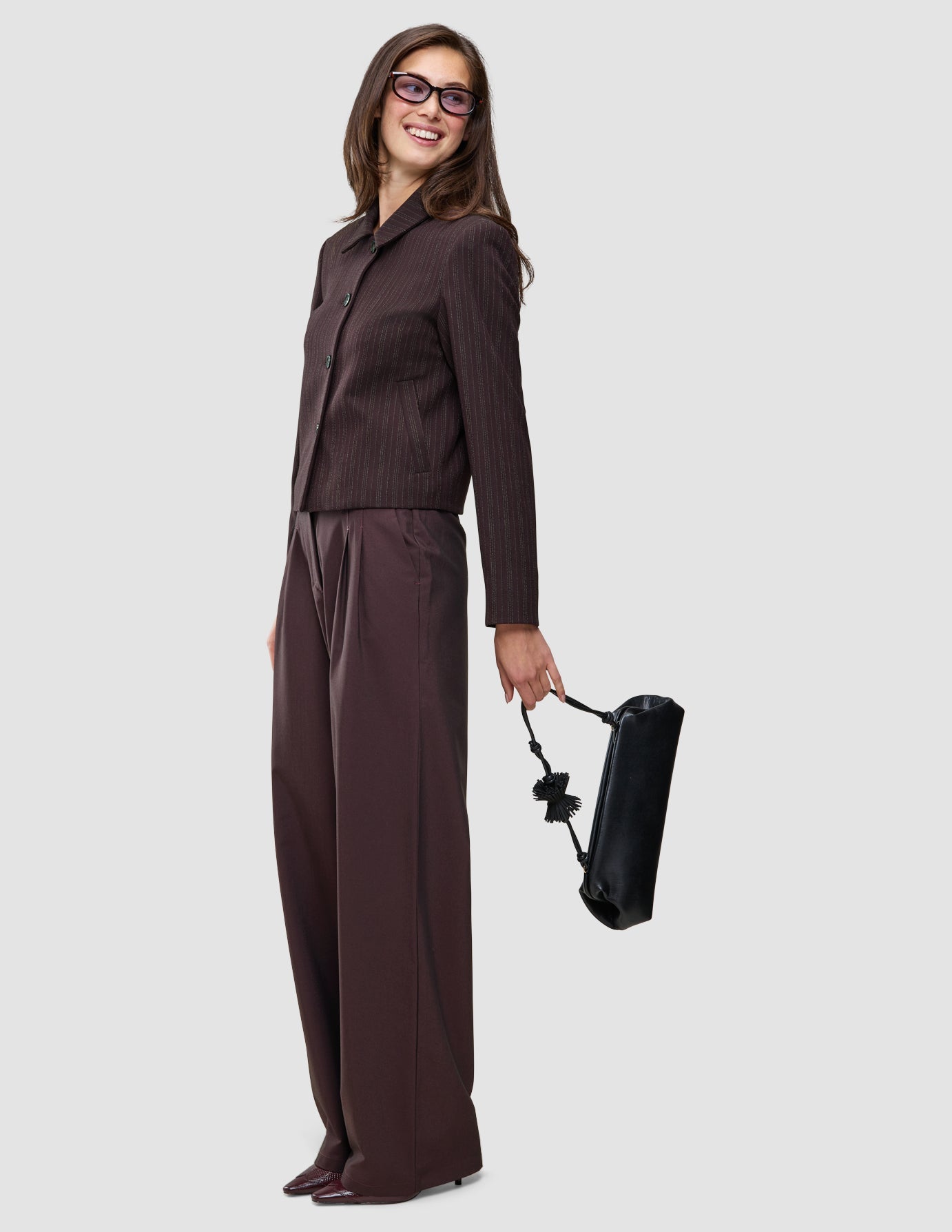 A smiling woman wearing a Burgundy from top to toe dark brown pinstripe jacket and wide-leg trousers, holding a black handbag.