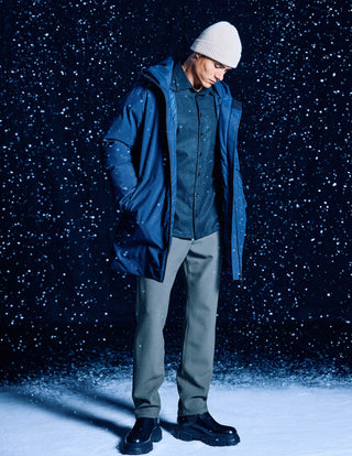 A person wearing a Dark Navy Utility Jacket over a button-down shirt, grey trousers, and a beanie, standing against a background of falling snow.