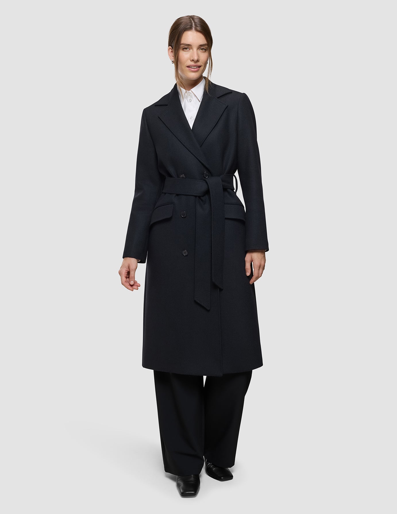 A person wears a Women’s Wool Coat Black, featuring a double-breasted design with a matching tie belt over a white collared shirt and black trousers, showcasing a clean Scandinavian style.