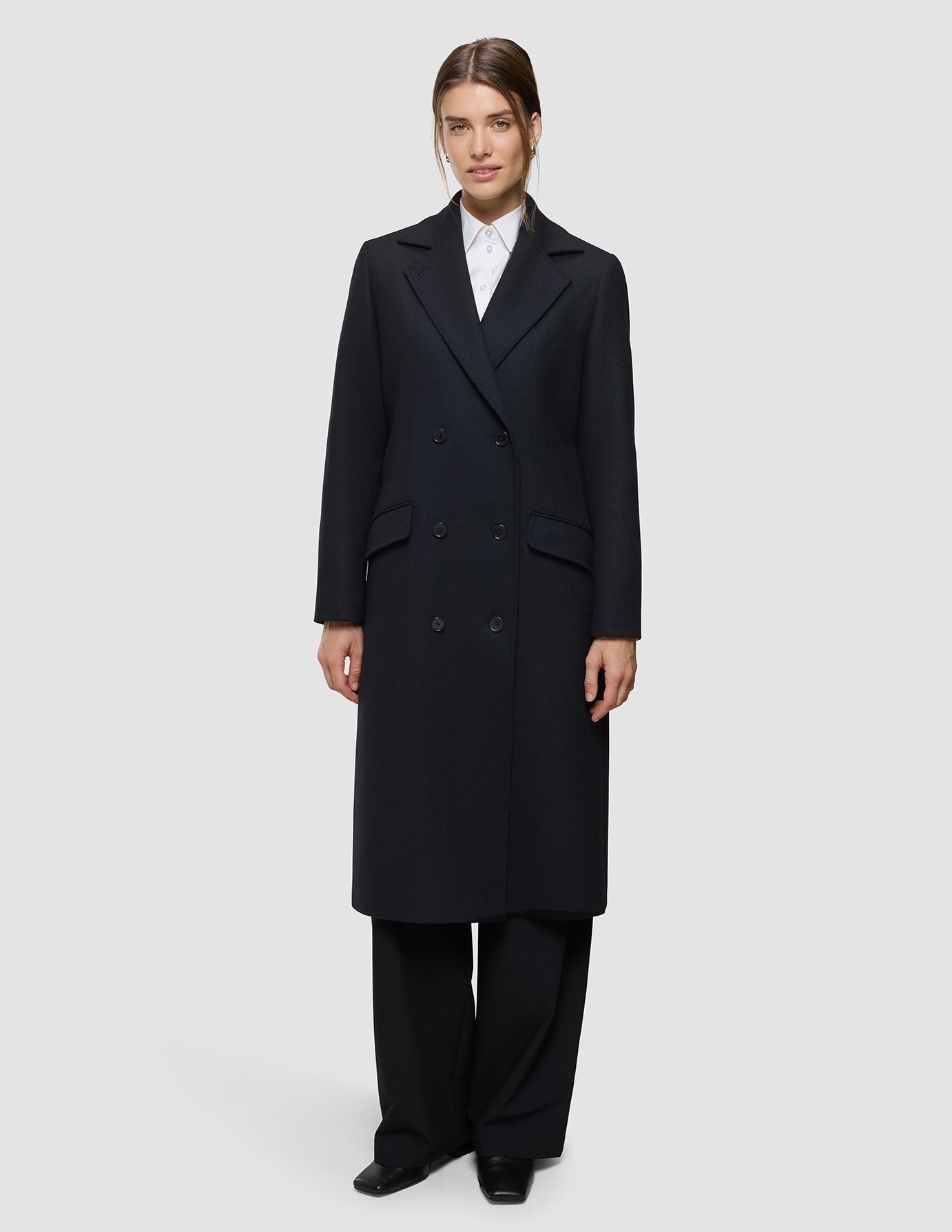 A woman wearing a Women’s Wool Coat Black in a Scandinavian design over a white collared shirt and black trousers.