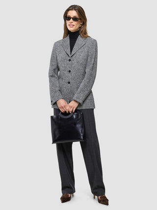 A woman wearing a Women Ashryn Blazer Domino Herringbone, black turtleneck, and dark trousers holds a black handbag.