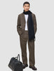 A man wearing a dark beige Atelier Overshirt heavyweight twill blazer over a white shirt with a dark scarf stands next to a black leather duffel bag.