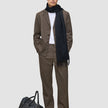 A man wearing a dark beige Atelier Overshirt heavyweight twill blazer over a white shirt with a dark scarf stands next to a black leather duffel bag.