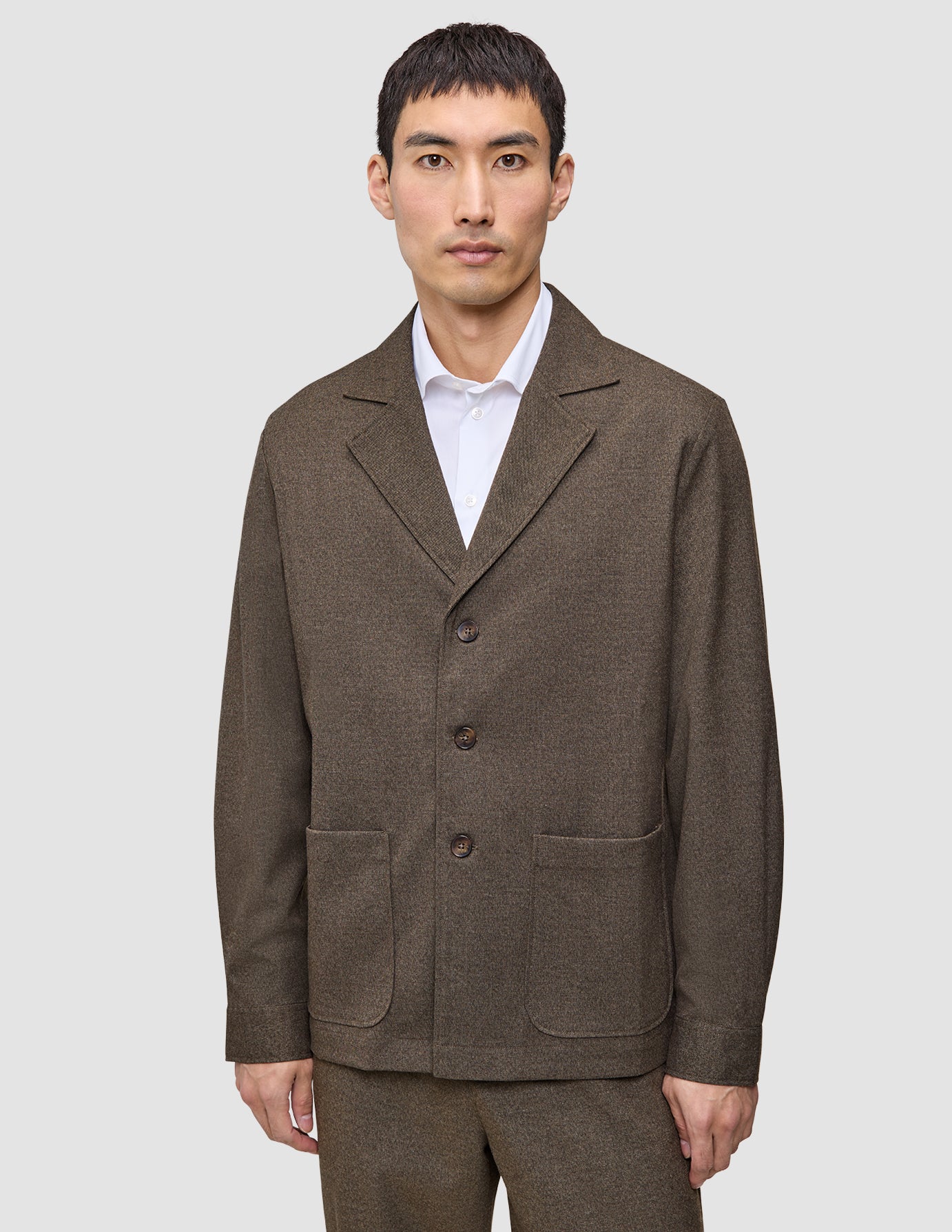 A man wears a brown Atelier Overshirt Dark Beige heavyweight twill blazer with large patch pockets over a white collared shirt.