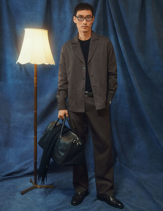 A man in a dark beige Atelier Overshirt, matching trousers, and a black shirt holds a dark leather duffel bag.
