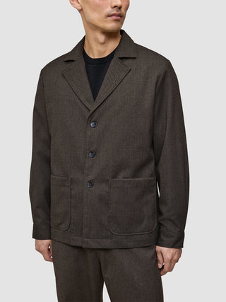A person wearing an Atelier Overshirt Dark Brown, a Scandinavian-designed blazer with notch lapels, three buttons, and two large patch pockets.