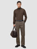 A man wears a brown quarter-zip sweater and Atelier Pants Relaxed Dark Beige in heavyweight broken twill, paired with a black belt, boots, and a brown duffel bag.