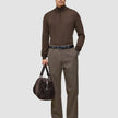 A man wears a brown quarter-zip sweater and Atelier Pants Relaxed Dark Beige in heavyweight broken twill, paired with a black belt, boots, and a brown duffel bag.