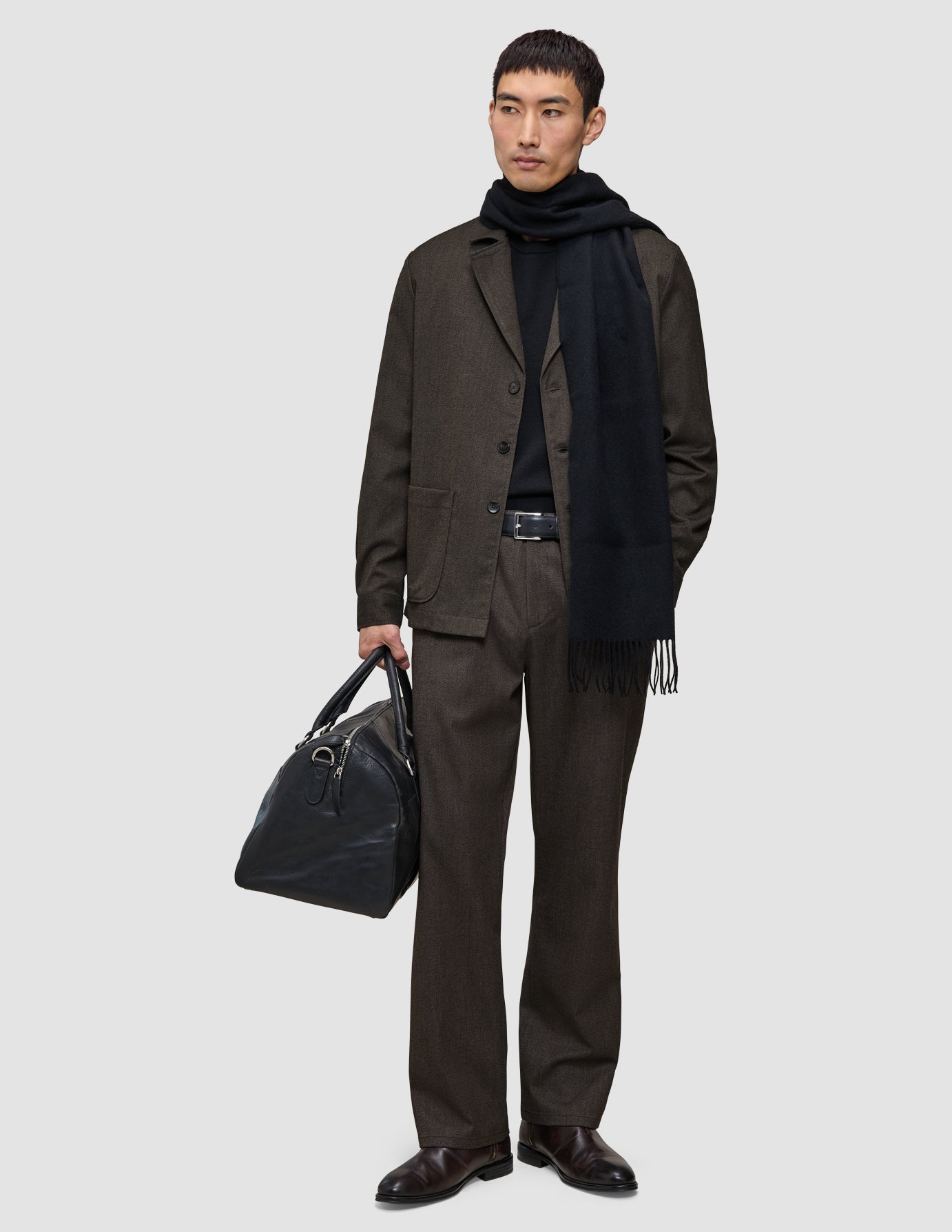 A man wearing an Atelier Pants Relaxed Dark Brown suit, a black sweater, and a scarf, while holding a black leather duffel bag.