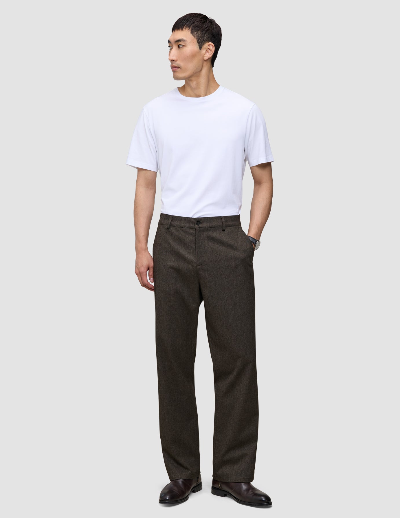 A person wearing a white t-shirt and Atelier Pants Relaxed Dark Brown in heavyweight broken twill, paired with dark leather shoes.