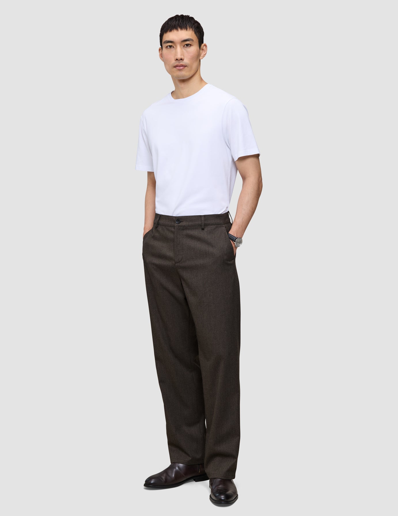 A man wearing a white t-shirt and Atelier Pants Relaxed Dark Brown in heavy-weight broken twill, standing with his hands in his pockets.