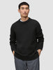 A man wears a black Chunky Soft Wool Knit Crewneck sweater with a thick ribbed collar and cuffs, with his hands in his pockets.