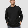 A man wears a black Chunky Soft Wool Knit Crewneck sweater with a thick ribbed collar and cuffs, with his hands in his pockets.