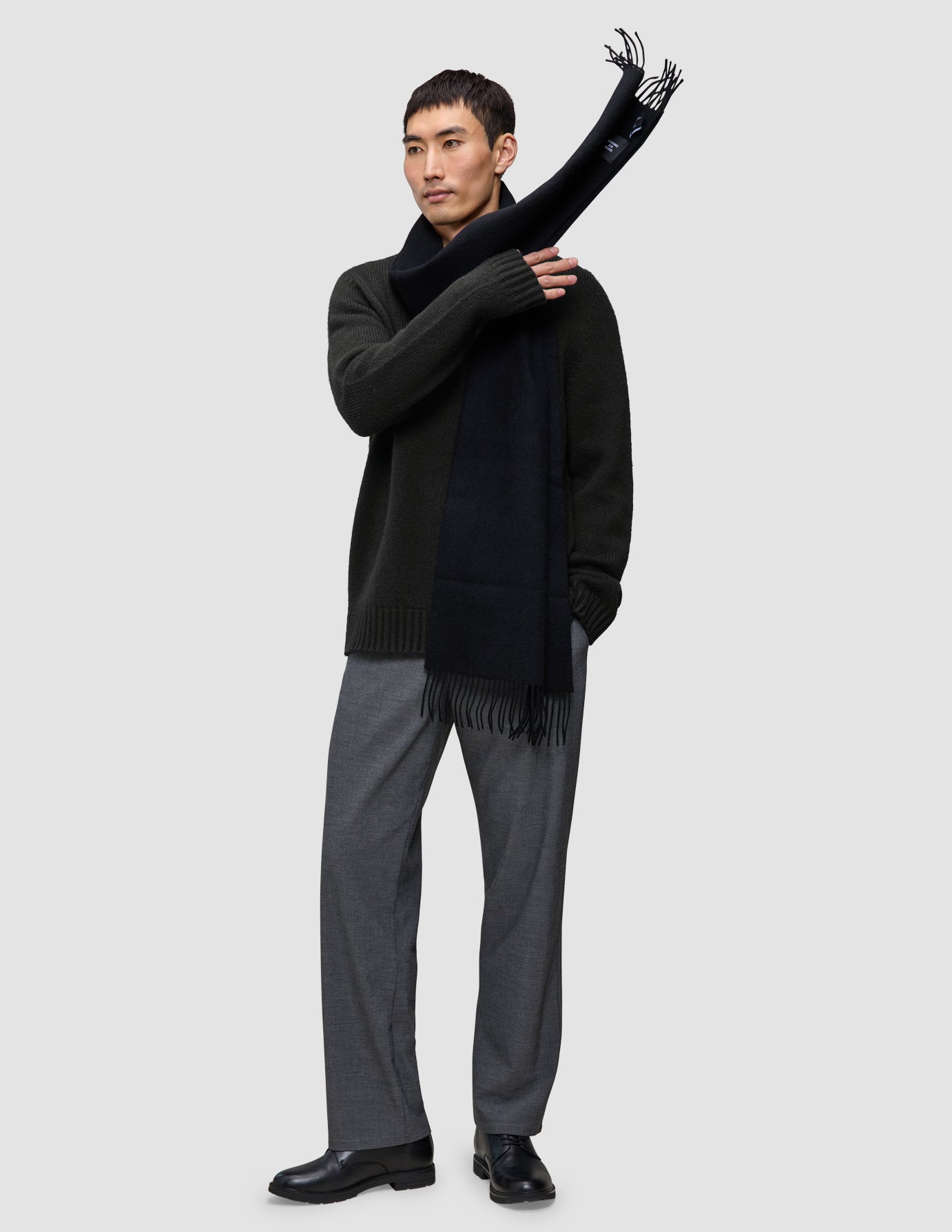 A man wearing a Chunky Soft Wool Knit Crewneck Black sweater in dark olive, grey trousers, and a black scarf draped over one shoulder.