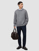 A man wearing a Chunky Soft Wool Knit Crewneck Glacier Grey over a white collared shirt, dark trousers, and dark loafers, holding a dark brown duffel bag.