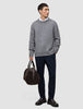 A man wears a Chunky Soft Wool Knit Crewneck Glacier Grey sweater over a white collared shirt, dark trousers, and dark loafers, while holding a brown duffel bag.