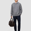 A man wears a Chunky Soft Wool Knit Crewneck Glacier Grey sweater over a white collared shirt, dark trousers, and dark loafers, while holding a brown duffel bag.