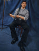 A man wearing a Chunky Soft Wool Knit Crewneck Glacier Grey sweater over a collared shirt, black trousers, and black shoes, sitting on a wooden chair.