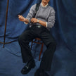 A man wearing a Chunky Soft Wool Knit Crewneck Glacier Grey sweater over a collared shirt, black trousers, and black shoes, sitting on a wooden chair.