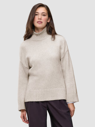 A woman wearing the Women’s Chunky Soft Wool Knit Turtleneck Sugar Melange, an oversized, light beige merino wool sweater with ribbed cuffs and hem in a minimalist Scandinavian design.
