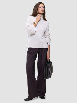 A woman wearing the Women’s Chunky Soft Wool Knit Turtleneck Sugar Melange, dark trousers, and glasses, while holding a black handbag.