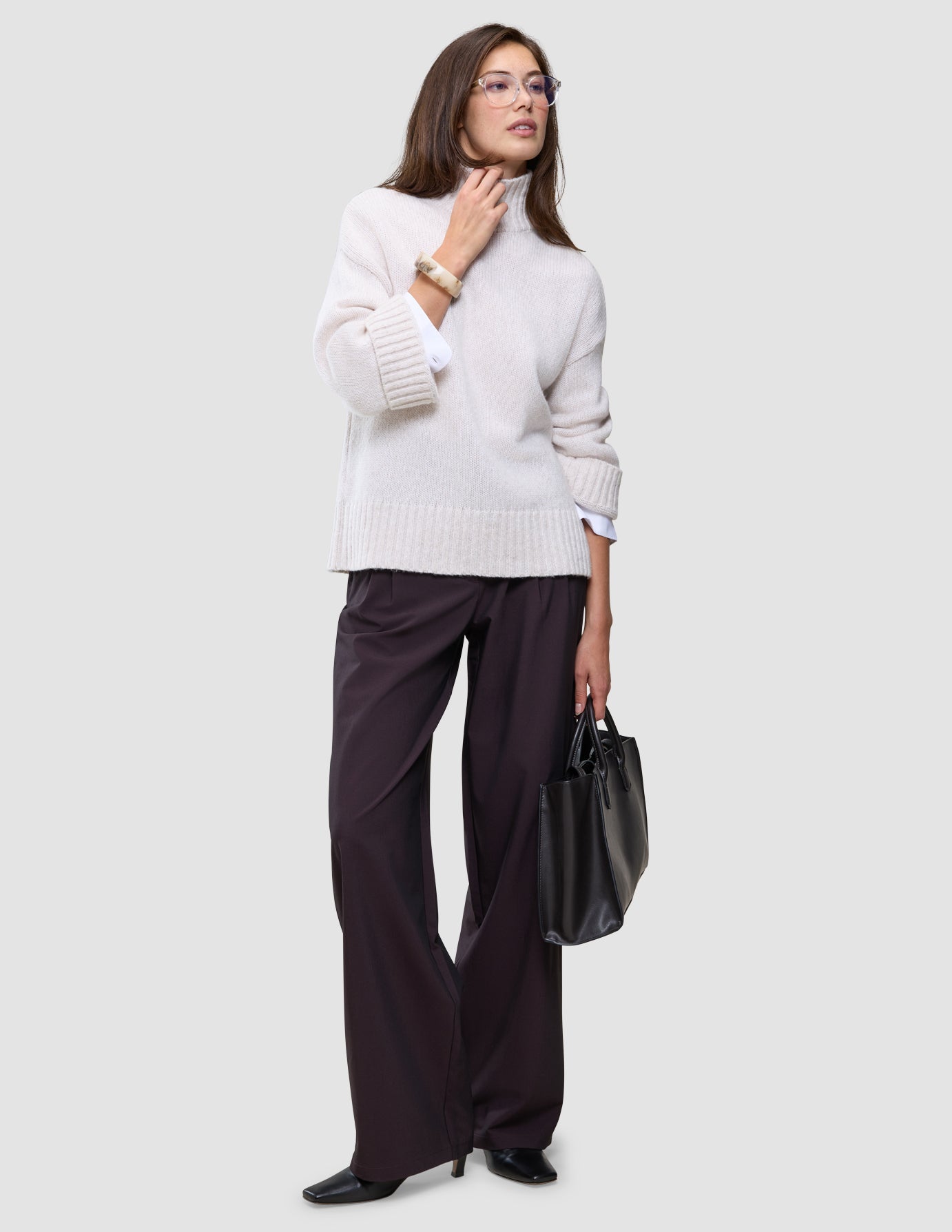 A woman wearing the Women’s Chunky Soft Wool Knit Turtleneck Sugar Melange, dark trousers, and glasses, while holding a black handbag.