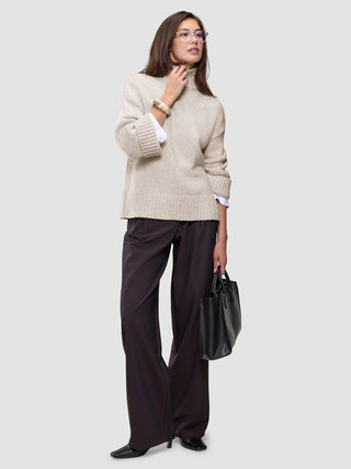 A woman wearing a Women’s Chunky Soft Wool Knit Turtleneck Sugar Melange in beige with wide-leg dark trousers, carrying a black handbag.
