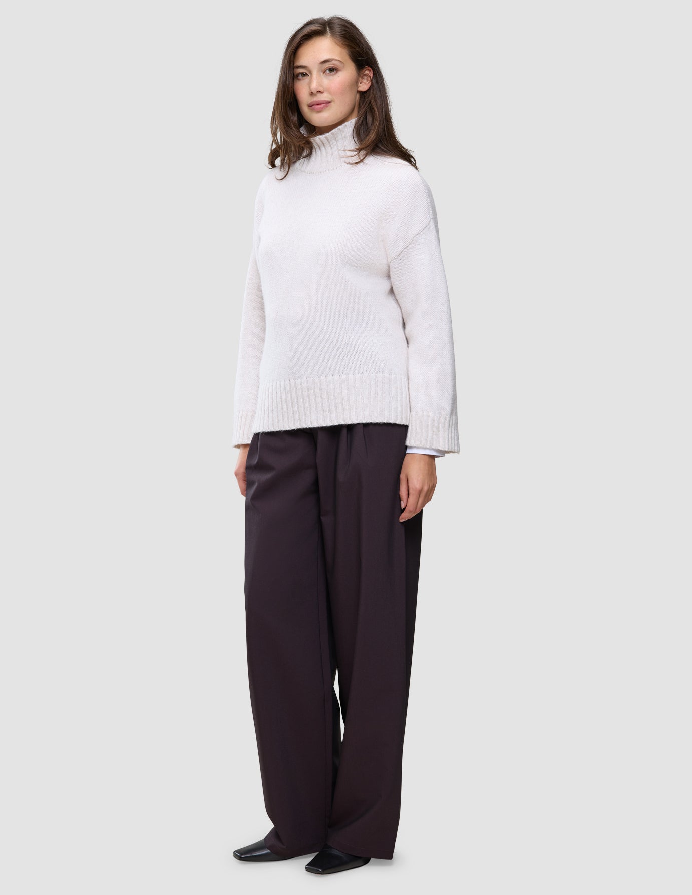 A woman wears an off-white Women’s Chunky Soft Wool Knit Turtleneck Sugar Melange and wide-leg dark trousers.