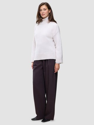 A woman wears an off-white Women’s Chunky Soft Wool Knit Turtleneck Sugar Melange and wide-leg dark trousers.