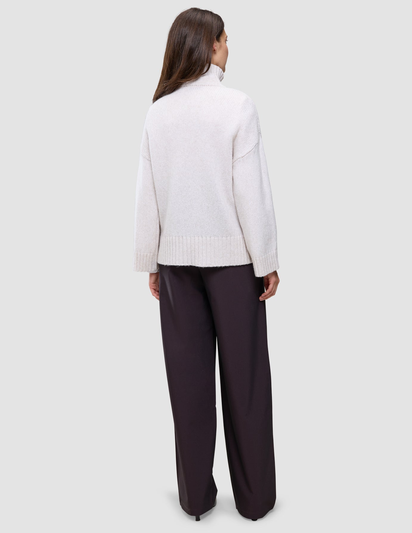 A person seen from behind, wearing an oversized, light-colored Women’s Chunky Soft Wool Knit Turtleneck Sugar Melange and wide-leg dark trousers.