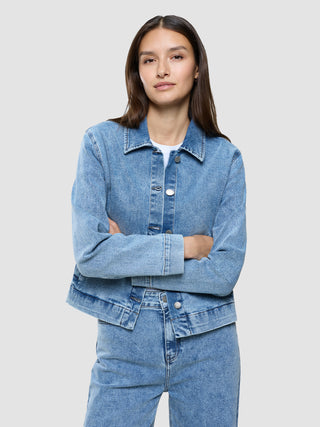 A woman with long brown hair wearing a Women’s Classic Denim Jacket Snow Blue Wash with her arms crossed.