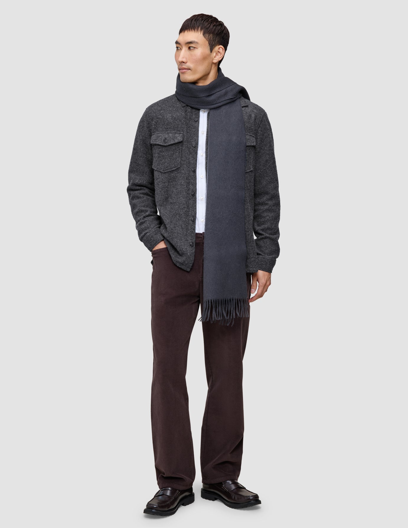 A man wearing a Double Layer Wool Overshirt Regular Dark Grey over a white shirt, a matching grey scarf, brown trousers, and dark loafers.