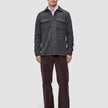 A man wearing a buttoned Double Layer Wool Overshirt Regular Dark Grey over a white collared shirt, paired with dark trousers and black loafers in a clean Scandinavian design.
