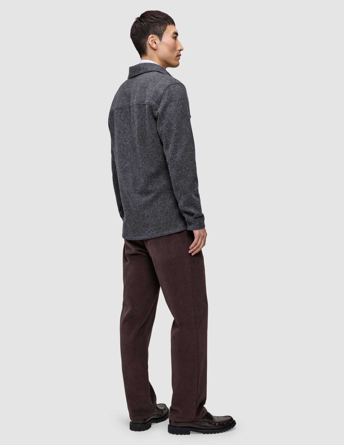 A man seen from the side wearing a Double Layer Wool Overshirt Regular Dark Grey, brown trousers, and dark loafers, showcasing a Scandinavian design aesthetic.
