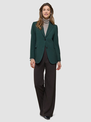 A woman wears a Women Ease Blazer Evergreen over a tan turtleneck paired with dark trousers.