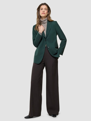 A woman wears a Women Ease Blazer Evergreen over a tan turtleneck paired with dark brown wide-leg trousers in a Scandinavian design.