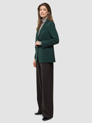 A woman wearing a Women Ease Blazer Evergreen over a grey turtleneck sweater and dark trousers.