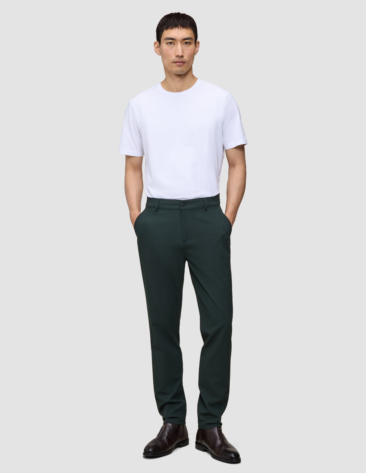 A man wearing a white t-shirt and dark green Ease Pants Regular Northern Pine with a tailored look, standing with his hands in his pockets.